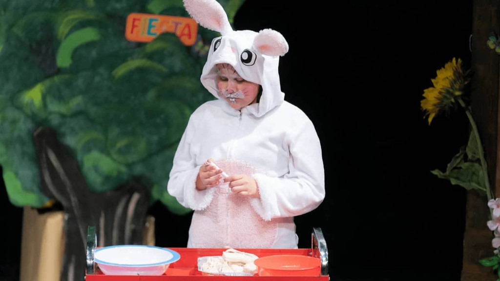 Young Cazateatro actor in a white rabbit costume performing on stage beside a Fiesta tree prop